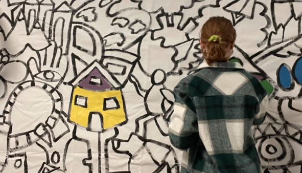 A student is standing in front of a mostly blank canvas filled with symbols and designs. The only painted item is a yellow house to the left of the student.